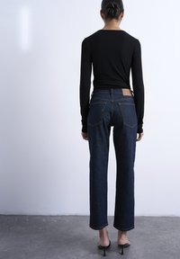 Femme debout face à un mur uni, portant un jean bleu foncé, un haut à manches longues noir et des sandales à talons noires, les cheveux attachés en un chignon bas.