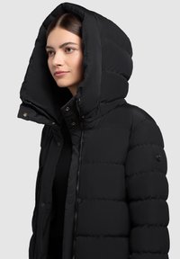 Jauna sieviete valkā melnu pūkainu jaku ar pārāk lielu kapuci, vērsta nedaudz pa kreisi uz viendabīga gaiši pelēka fona.