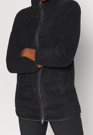 Personne portant une veste polaire zippée noire texturée, les bras croisés, montrée de la poitrine jusqu'aux hanches, sur un fond uni.