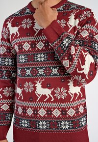 Mann trägt einen roten Weihnachtspullover mit weißen Rentier- und Schneeflockenmuster, Hand berührt den Kragen.