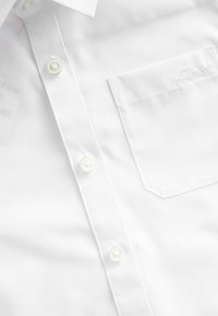 Chemise blanche à manches longues en tissu lisse, dotée d'une poche avec logo brodé et de boutons ressemblant à des perles à l'avant.