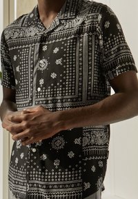 Chemise noire à manches courtes avec motifs paisley blancs et pois, dotée d'un col cranté et d'une patte de boutonnage. Texture de tissu douce.