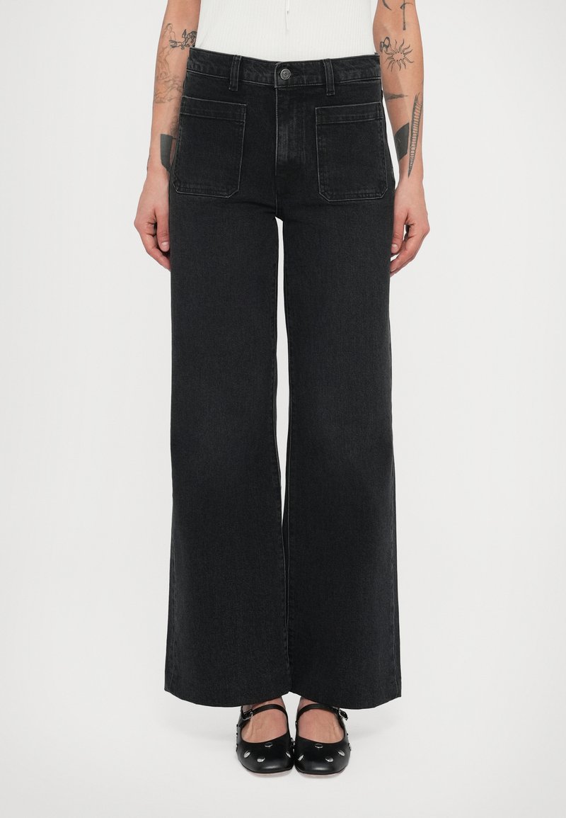 Sieviete valkā melnas platu kāju augsta vidukļa džinsas ar priekšējām uzšuvuma kabatām un melnas Meri Džeina kurpes ar sudraba kniedēm.