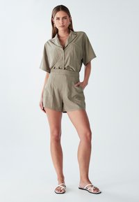 Moteris stovi su ranka kišenėje, vilkinti smėlio spalvos trumpomis rankovėmis marškinėlius su saga ir suderintus aukštai užtraukiamus šortus bei baltus šlepetes.