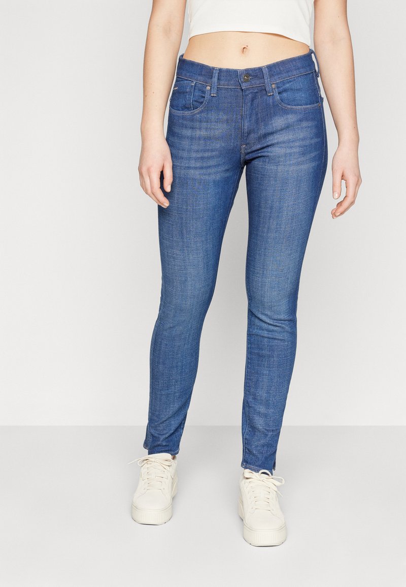 Smal jeans i mellomblå denim, med knappelukking, beltehemper og frontlommer. Brukt med hvite sneakers.