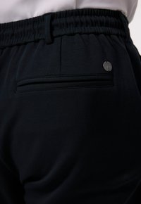 Detailní záběr na černé elastické kalhoty s pasem, vzadu s kapsou s klopou a malou kulatou kovovou štítkovou nálepkou blízko kapsy.