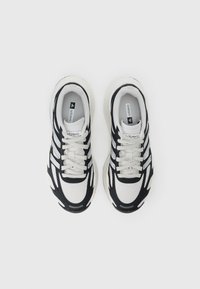Tênis atléticos em preto e branco com parte superior em malha, detalhes texturizados e sola acolchoada, apresentando riscas e atacadores em contraste.