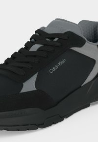Μαύρο και γκρι αθλητικό παπούτσι Calvin Klein με μαύρα κορδόνια, επενδεδυμένο γιακά και λογότυπο στο πλάι και στη γλώσσα, σε κοντινή λήψη.