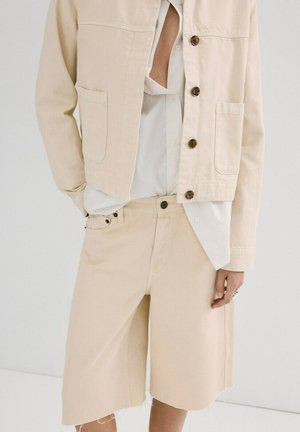 Persona con chaqueta beige abotonada, camisa blanca parcialmente metida y pantalones cortos beige a la rodilla a juego con dobladillos deshilachados.