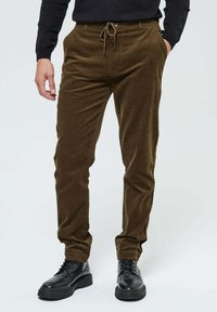 Pantalon en velours côtelé marron foncé avec une coupe fuselée, taille élastique avec cordon de serrage, poches latérales, porté avec des chaussures noires à lacets.