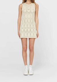 Krémové pletené šaty s geometrickými výřezy, bez rukávů a s vlnitým lemem. Vydané s bílými vysokými teniskami.
