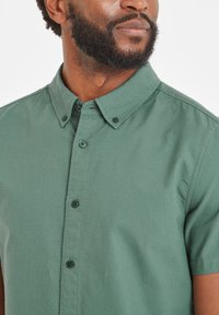 Short-sleeve button-up shirt in dark green cotton, featuring a classic collar, front buttons, and a smooth, textured fabric.