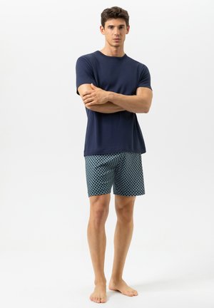 Jonge man staat op blote voeten, draagt een marineblauw shirt met korte mouwen en blauw-witte korte broek met patroon, armen gekruist, neutrale achtergrond.