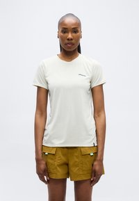 Γυναίκα με τα μαλλιά δεμένα πίσω που φοράει ένα απλό ανοιχτό μπεζ T-shirt Patagonia και μουσταρδί σορτς με μπροστινές τσέπες, στεκόμενη μπροστά σε λευκό φόντο.