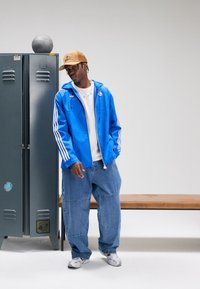 Blå nylonjacka med vita ränder, vit t-shirt, oversize denimjeans och silvriga sneakers. En beige keps med ett texturerat mönster används.