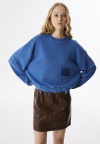 Blauwe oversized sweatshirt met ribbelmanchetten en -zoom, met een zwart tekstlogo. Gecombineerd met een bruine leren minirok met een gladde textuur.