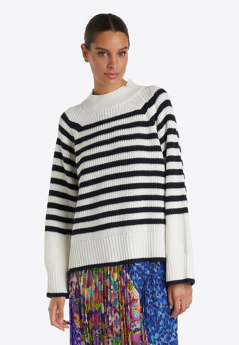 Frau in einem weiß und schwarz gestreiften Strickpullover, kombiniert mit einem bunten Blumenkleid und einem blauen gemusterten Rock vor einem weißen Hintergrund.