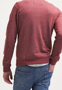 Ciemnoczerwona teksturowana bluza z długimi rękawami i ściągaczami na mankietach, zestawiona z niebieskimi dżinsami z tylnymi kieszeniami.