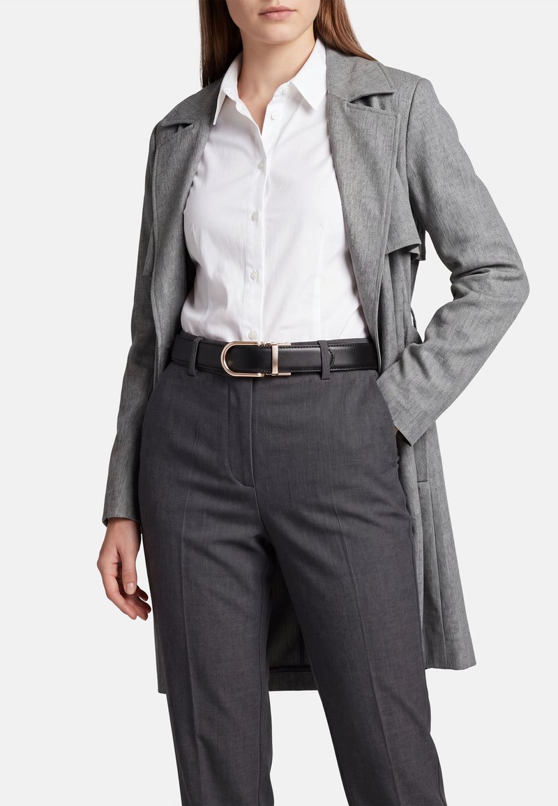 Donna che indossa una camicia bianca con bottoni, pantaloni grigi su misura con cintura nera e un lungo cappotto grigio, in piedi con una mano in tasca.