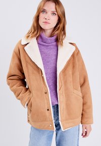 Giacca in suede sintetico marrone con colletto in pile bianco, dotata di chiusura con bottoni, due tasche frontali e una vestibilità morbida. Indossata sopra un dolcevita viola.