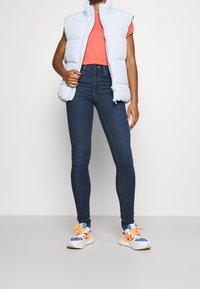 Chaleco acolchado sin mangas de color azul claro sobre una camiseta de color coral, combinado con jeans ajustados de color azul oscuro y zapatillas deportivas de gran tamaño multicolores.