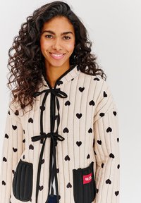 Béžová prešívaná bunda s dlhými rukávmi, s vertikálnymi čiernymi pruhmi a vzorom srdiečok. Obsahuje čierne viazanie na zapínanie a dva akcenty v podobe vreciek.