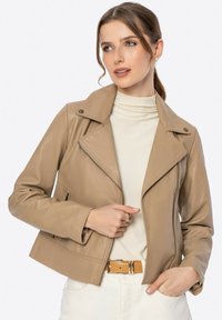 Hellbeige Bikerjacke aus Leder mit einem Reverskragen, schmaler Passform und seitlichen Reißverschlusstaschen; getragen über einem cremefarbenen Rollkragenpullover. Glatte Textur.