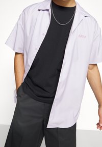 Camicia a maniche corte button-up di colore lavanda con una vestibilità rilassata, indossata sopra una T-shirt nera, caratterizzata da una leggera texture e dettagli ricamati sottili.