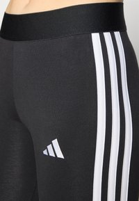 Juodos sportinės legging'os su plačiu juodu juosmeniu, kuriuose yra trys baltos vertikalios juostelės išorinėje pusėje ir baltas logotipas.