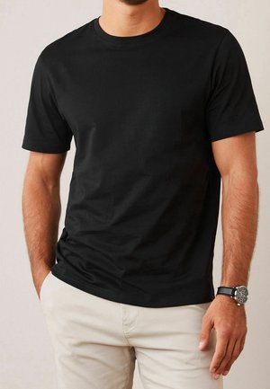 Homme portant un t-shirt noir uni à manches courtes, un pantalon beige et une montre-bracelet noire avec un cadran rond, debout avec une main dans la poche.