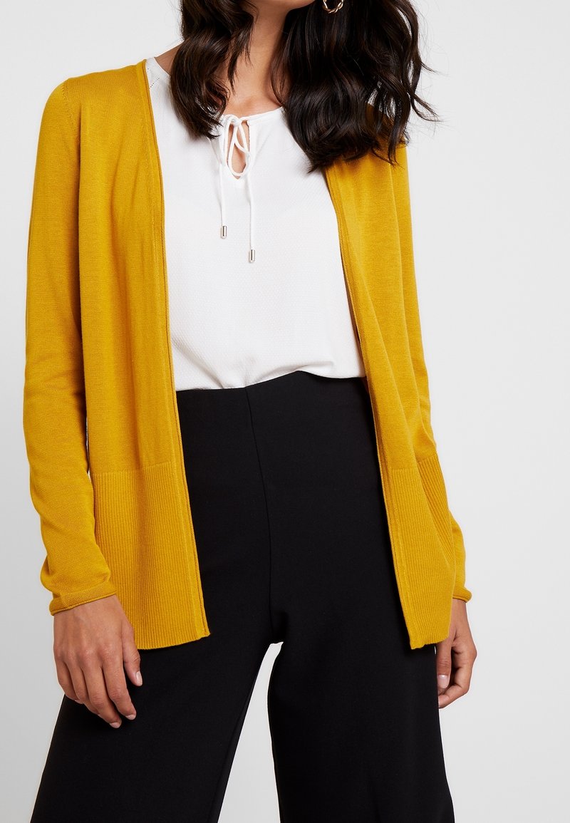 Femme portant un cardigan ouvert jaune moutarde, un chemisier blanc avec des liens à l'avant, et un pantalon noir taille haute sur un fond clair.