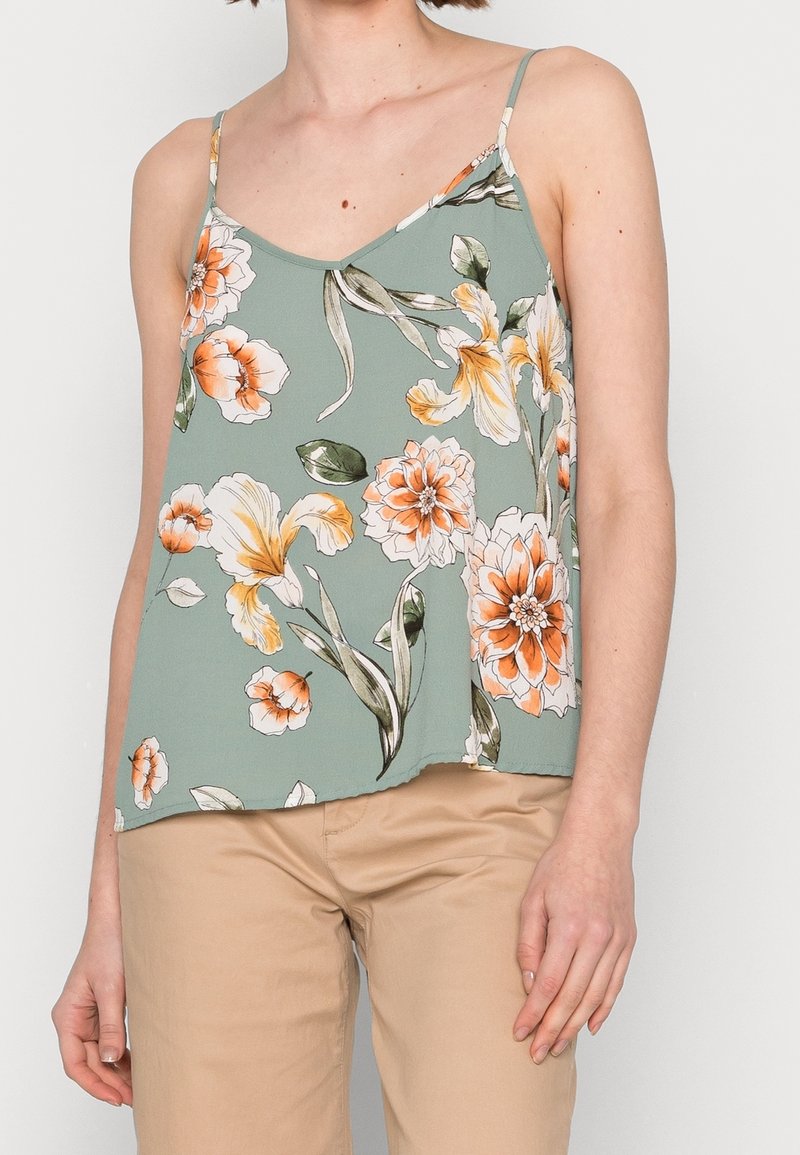 Femme portant un caraco vert clair avec un imprimé floral orange et blanc et un pantalon beige sur un fond uni.