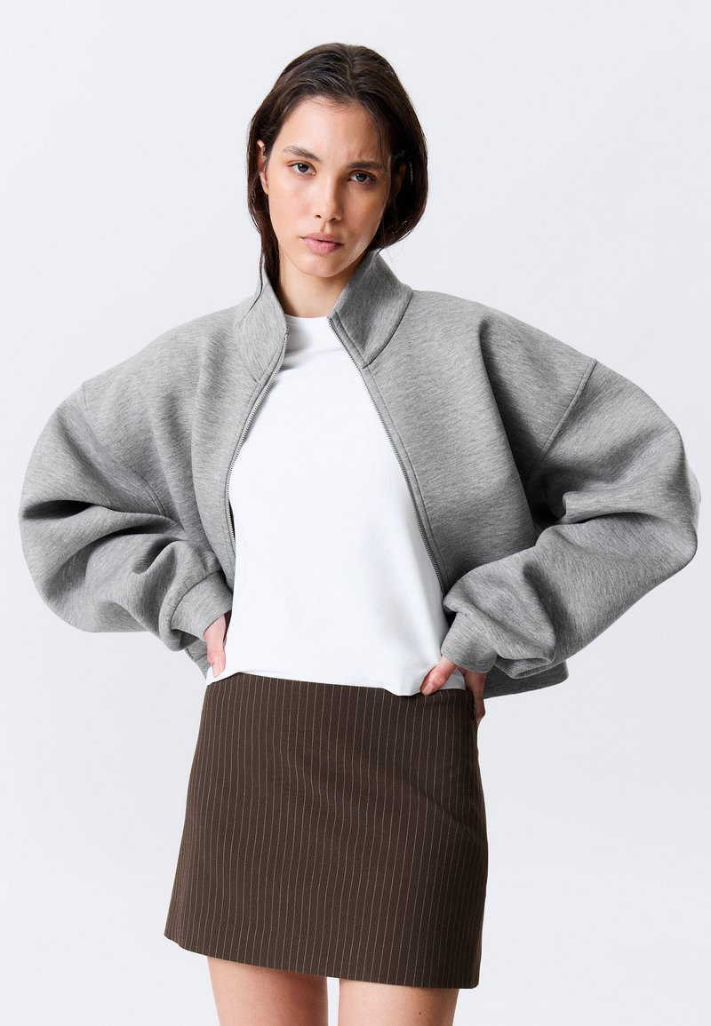 Sudadera gris de gran tamaño con cremallera y mangas abullonadas, combinada con una camiseta blanca y una minifalda marrón con rayas diplomáticas.