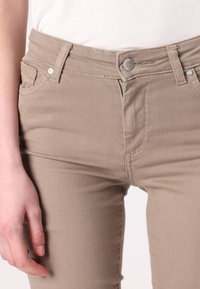 Beige korkeavyötäröiset farkut, joissa on vetoketju ja nappikiinnitys. Ominaisuuksiin kuuluu kaksi etutaskua, sileä pinta ja näkyvät saumat.