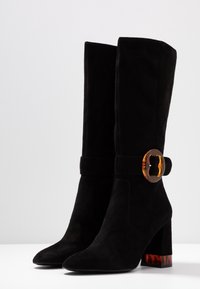 Botas de ante negras hasta la rodilla con un tacón grueso y una hebilla de acento de carey. Textura suave, punta afilada y construcción robusta.