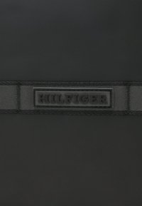 Sort struktureret stof med et glat sort gummipatch, der har "HILFIGER" prægede i midten, omkranset af et vævet sort bånd.