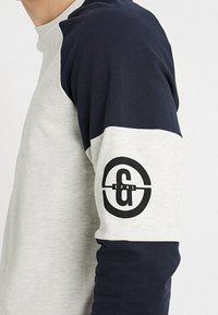 Camisa de manga larga hecha de una mezcla de algodón; presenta un cuerpo gris claro, mangas azul marino y un prominente logo circular negro en el brazo.