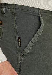 Olivegrüne Baumwollhose mit glatter Textur, Seitentasche, kontrastierenden Nähten und einem kleinen schwarz-gelben Markenetikett.