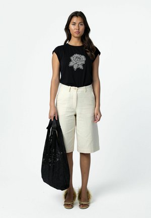 Woman wearing black sleeveless top with white rose design, cream knee-length shorts, holding large black bag, and fuzzy open-toe sandals.