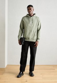 Sweat à capuche vert olive avec logo blanc, poche kangourou et cordon de serrage. Porté avec un pantalon noir à jambes droites et des chaussures à lacets noires.