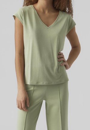 Frau trägt ein hellgrünes, kurzärmeliges Oberteil mit V-Ausschnitt und passende Hose, steht mit einer Hand in der Hüfte vor einfarbigem Hintergrund.