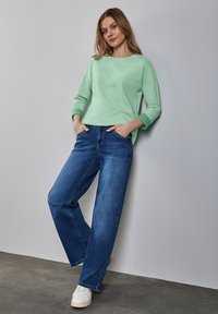 Ljusgrön långärmad tröja med ribbade ärmslut parat med vida blå jeans. Modellen står mot en neutral vägg, iförd vita sneakers.