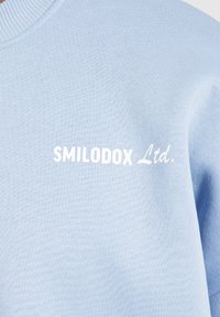 Lichtblauwe sweatshirt met geribde kraag, voorzien van witte tekst "SMILODOX Ltd." op de voorkant. Zachte textuur, minimalistisch ontwerp.