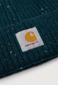 Tessuto lavorato a maglia verde scuro con macchie multicolori, caratterizzato da un'etichetta quadrata cucita di colore bianco con un logo giallo e il testo "Carhartt" in rosso.