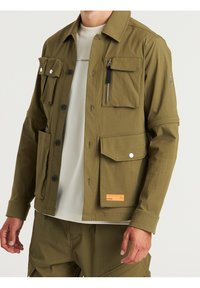 Mężczyzna w oliwkowej kurtce zapinanej na guziki z wieloma kieszeniami, na białej koszulce z okrągłym dekoltem i pasujących oliwkowych spodniach.