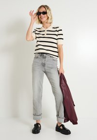 Mujer con gafas de sol negras, camiseta de rayas de manga corta, jeans grises con dobladillo, zapatos negros, sosteniendo una chaqueta burdeos frente a un fondo blanco liso.