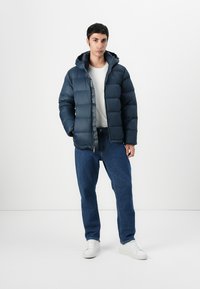 Navy puffer jacket with a hood, zipped front, and quilted texture. Paired with light jeans and white sneakers.