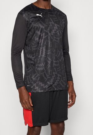 Mann trägt schwarzes Sportshirt mit langen Ärmeln und abstraktem Muster sowie schwarze Shorts mit rotem Einsatz an der Seite, steht vor einfarbigem Hintergrund.