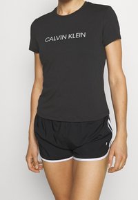 Camiseta de algodón negra con mangas cortas que presenta el logo "CALVIN KLEIN" en blanco. Combinada con pantalones cortos deportivos negros con acentos blancos.