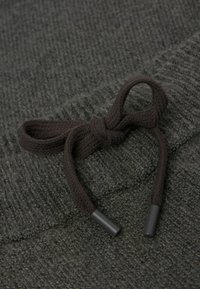 Tmavě šedá pletená látka s uvázanou tmavě hnědou šňůrkou se hladkými plastovými konci, umístěna na texturovaném pozadí.